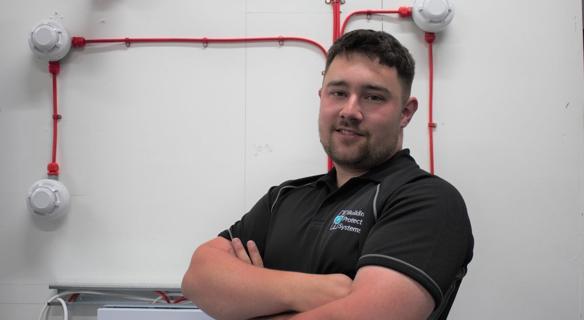 Young man with dark hair wearing short sleeved polo shirt with work branding standing in front of alarm testing board